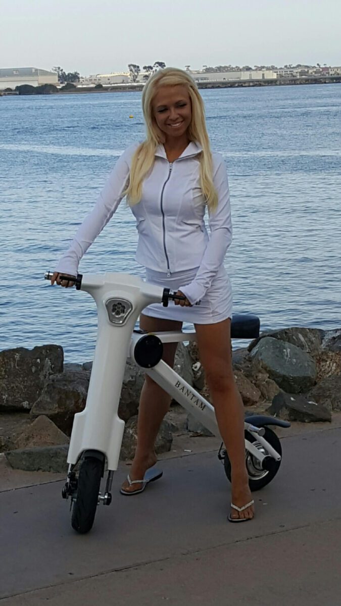 A blond woman posing next to a white electric scooter.